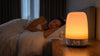 Sunrise alarm clock glowing warm on a bedside table beside a sleeping woman